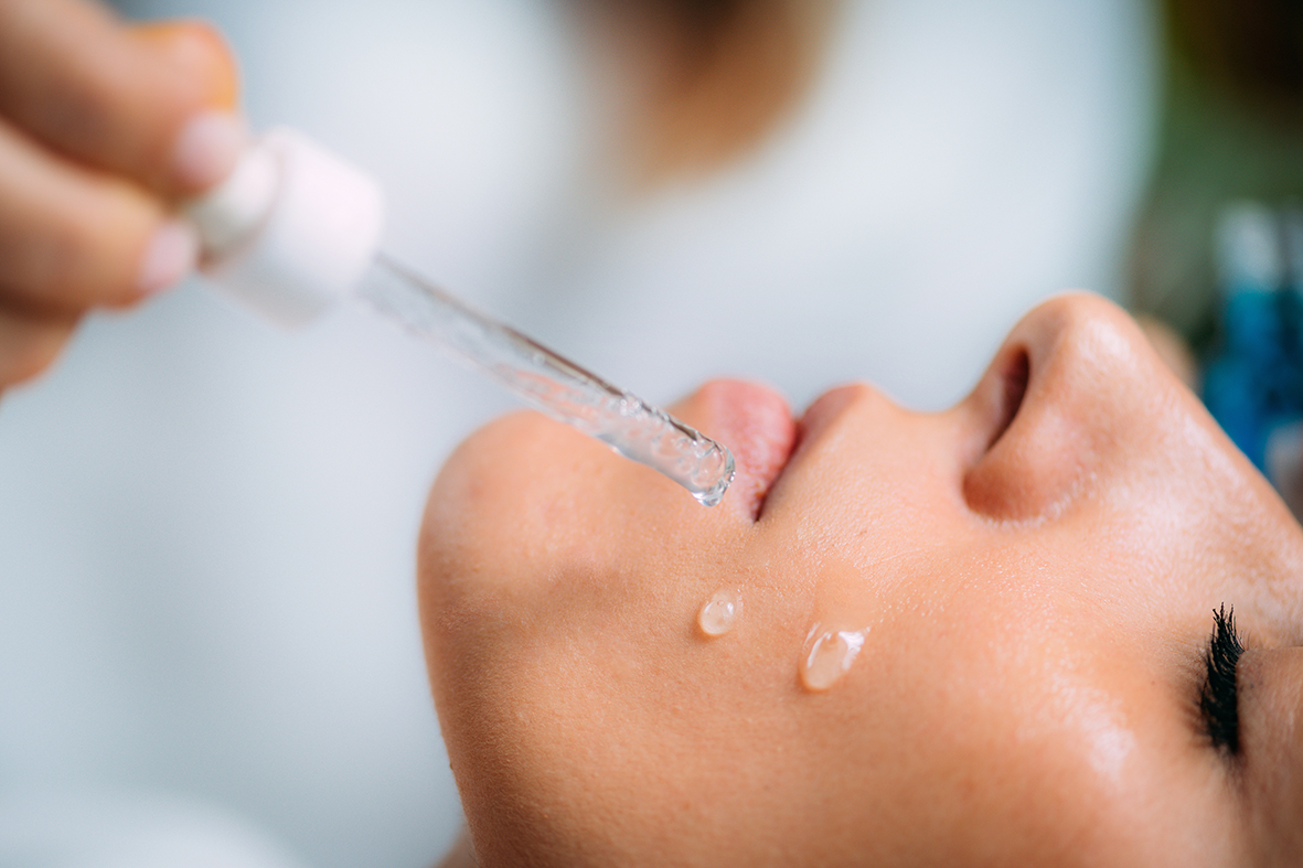A beautician is dripping hyaluronic acid from a dropper onto a woman’s face.